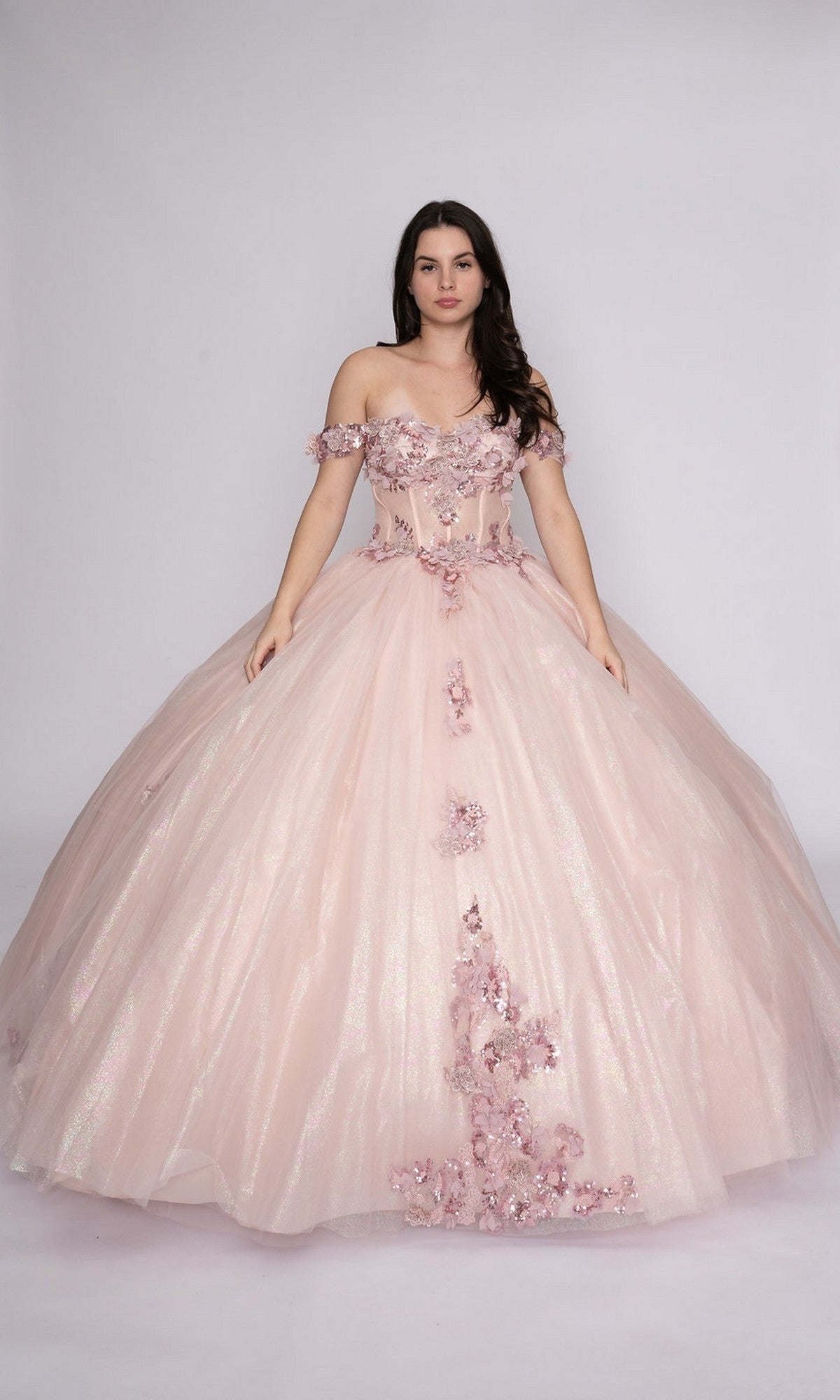 A woman models the blush Quinceanera Dress Q116 by Nina Canacci, featuring an off-the-shoulder design with floral embellishments and a full tulle skirt, standing against a plain background.